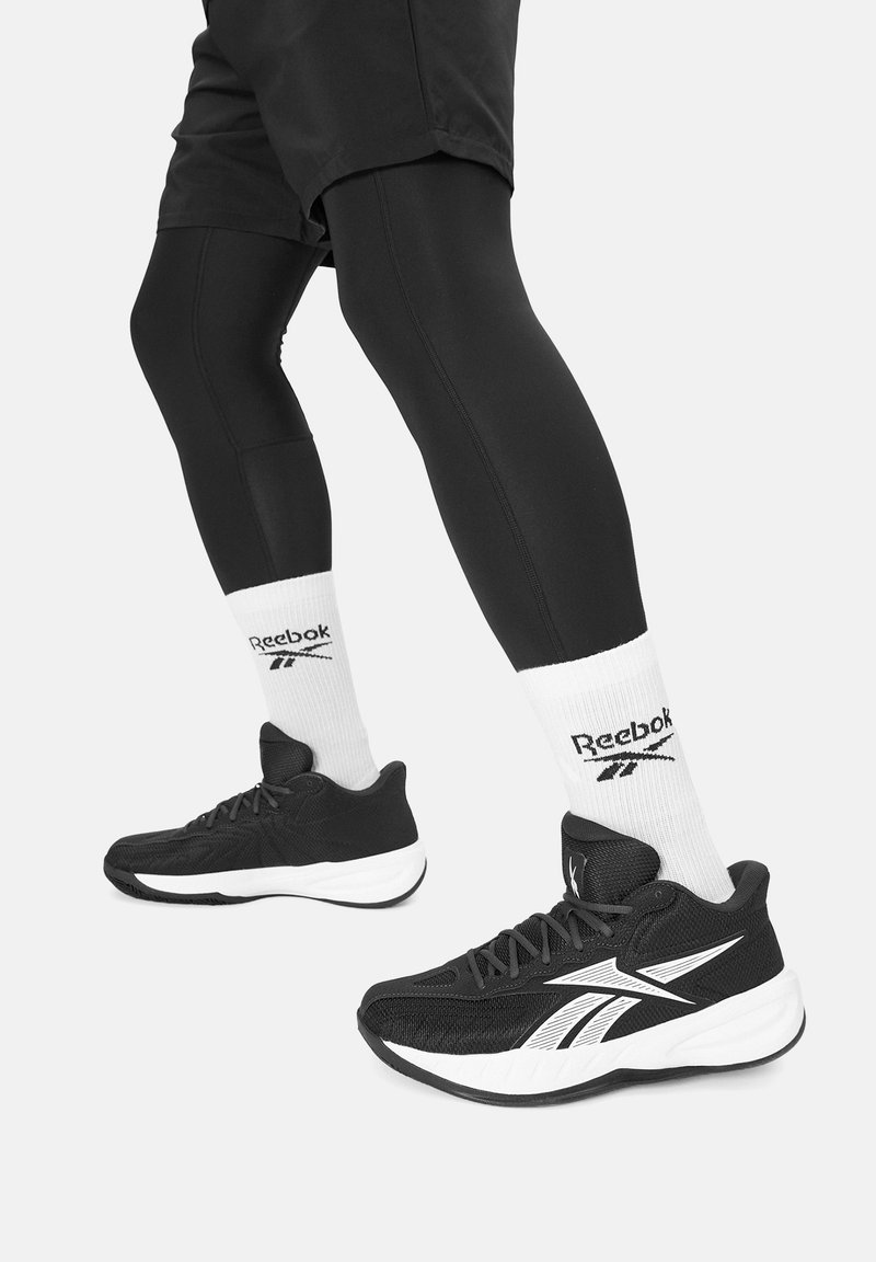Czarne buty sportowe z białymi akcentami, zestawione z czarnymi legginsami i białymi skarpetkami z logo Reebok. Buty mają teksturowaną powierzchnię.