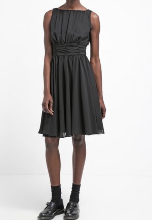 Czarna sukienka bez rękawów, sięgająca do kolan, z marszczonym detalem w talii, noszona z czarnymi skarpetkami i czarnymi butami z klamrą na ciemnoskórej osobie.