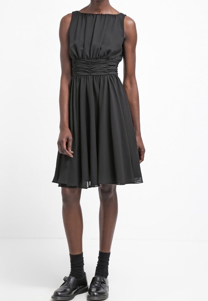 Robe noire sans manches jusqu'aux genoux avec un détail de taille froncée, portée avec des chaussettes noires et des chaussures à boucle noires sur une personne à la peau foncée.