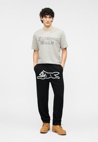 Γκρι βαμβακερό T-shirt με το σχέδιο «Icecream World», συνδυασμένο με μαύρο παντελόνι φόρμας που φέρει λευκό σχέδιο καρτούν σκύλου και καφέ μπότες.