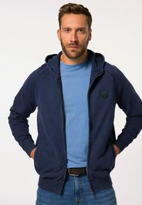 Tmavě modrá mikina na zip s kapucí, žebrovanými manžetami a předními kapsami. Na hrudi má strukturované logo. Nosená přes světle modrou košili.