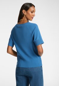 Stickad blå tröja med korta ärmar, ribbad struktur, rund halsringning och en liten emblemdetalj bak. I kombination med jeanstroser.