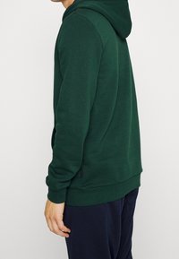 Felpa con cappuccio verde scuro, dal texture morbido, con maniche lunghe, tasca a marsupio e cappuccio regolabile con cordino. Presenta un orlo e polsini a costine.