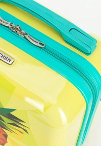 Maleta rígida amarilla con asa y cremallera turquesa, con un gráfico de piña y una placa metálica de la marca.