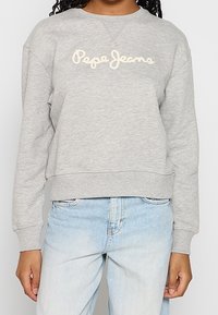 Sudadera gris recortada con borde acanalado, con el logotipo bordado de "Pepe Jeans" en color blanco hueso. Combinada con pantalones vaqueros azul claro.