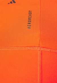 Urheilullinen polyesterikangas kirkkaassa oranssissa värissä, jossa on hienoa ribbiä. Varustettu mustalla "AEROREADY" -merkillä ja näkyvillä sauman yksityiskohdilla.