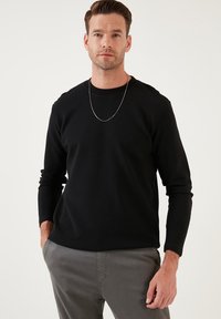 Fekete, hosszú ujjú póló bordázott textúrával, kerek nyakkivágással és ezüst lánc nyaklánccal, világosszürke nadrággal párosítva.