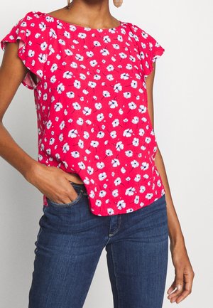 Vrouw draagt een rode korte mouwen blouse met witte bloemenprint en donkerblauwe spijkerbroek, hand nonchalant in de zak, tegen een effen achtergrond.