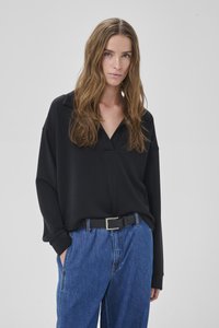 Donna con lunghi capelli castani mossi, indossa una maglietta nera a scollo a V morbida infilata in jeans blu a vita alta con cintura nera, in piedi con le mani nelle tasche.