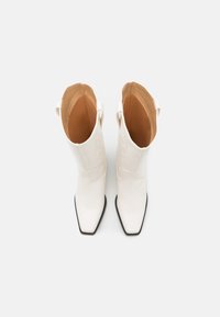 Botas de vaquero de cuero blanco con punta puntiaguda, tacón apilado y interior marrón. Costuras decorativas a lo largo de los lados exteriores.