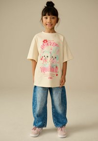 Korte mouw crèmekleurig t-shirt met kleurrijke konijnafbeelding en tekst, gecombineerd met wijde blauwe denim jeans en lichtroze sneakers.