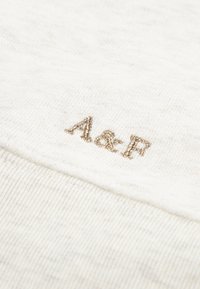 Biely textil v detaili s vyšívanými písmenami "A&F" zlatou niťou. Materiál má jemnú textúru s jemnými, svetlými bodkami.