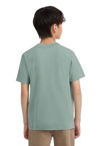 Deček stoji obrnjeno, oblečen v enostaven svetlo zelen kratek rokav majico in bež hlače, na beli podlagi.