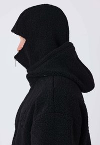 Personne en veste noire texturée à capuche avec fermeture éclair couvrant le bas du visage, présentée de profil sur un fond clair uni.