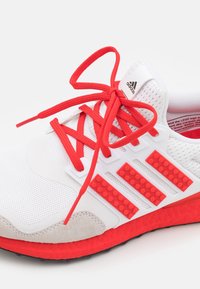 Bílé tenisky s červenými akcenty, síťovaným vrškem, texturovanou gumovou podrážkou a výrazným logem se třemi pruhy s detailním zpracováním připomínajícím LEGO. Červené tkaničky.