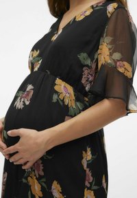 Zwarte bloemenjurk met korte, luchtige mouwen; heeft grote gele en roze bloemen en een gather taille, die een ronde buik accentueert.