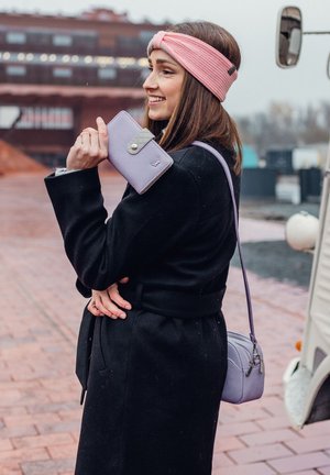 Light purple wallet with a textured flap and snap closure held in one hand, paired with a small crossbody bag. Black coat, pink headband.