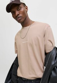 Lichtbeige katoenen t-shirt met een subtiel logo, gecombineerd met een zwarte leren jas, een zilveren ketting en een bruine pet.