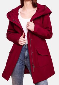Veste imperméable rouge avec fermeture éclair et boutons-pression, grandes poches avant, capuche et finition en tissu texturé. Idéale pour les activités en extérieur.