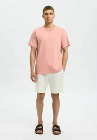 Rosa bomullströja med rund hals, i kombination med ljusbeiga linne-shorts. Modellen bär svarta sandaler med dubbla remmar.