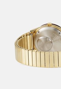 Montre en acier inoxydable doré avec un bracelet poli et un fond complexe orné de détails gravés. Boîtier rond et couronne texturée.