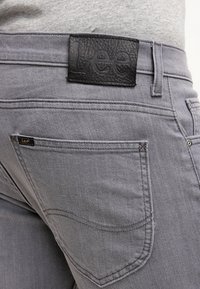 Vaqueros de mezclilla gris con una etiqueta de cuero negro texturizado en la parte posterior de la cintura. Las características incluyen dos bolsillos traseros y costuras en contraste.