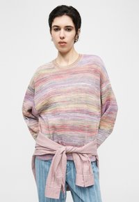 Person, die einen mehrfarbigen pastellfarbenen Strickpullover trägt, mit einer rosa strukturierten Jacke, die um die Taille gebunden ist, und blauen gestreiften Jeans, vor einem schlichten Hintergrund.