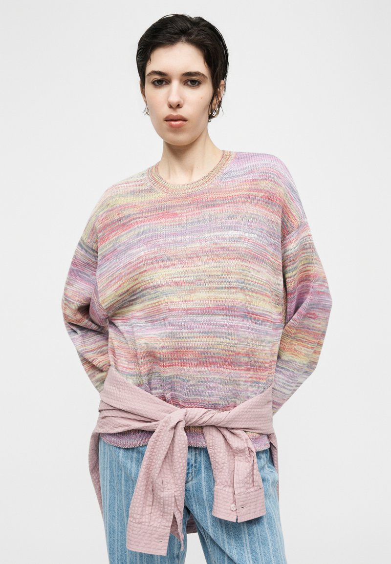 Persona che indossa un maglione lavorato a maglia dai colori pastello multicolori con una giacca rosa testurizzata legata in vita e jeans a righe blu, su uno sfondo semplice.