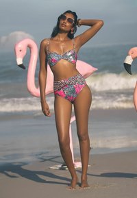 Cvečni bikini v modri, roza in črni barvi z visokim pasom. Model pozira blizu roza flamingov na plaži z morskimi valovi v ozadju.