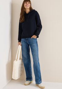 Felpa con cappuccio blu navy, fit rilassato, abbinata a pantaloni larghi in denim azzurro. Una borsa tote bianca e sneakers color crema completano l'outfit.