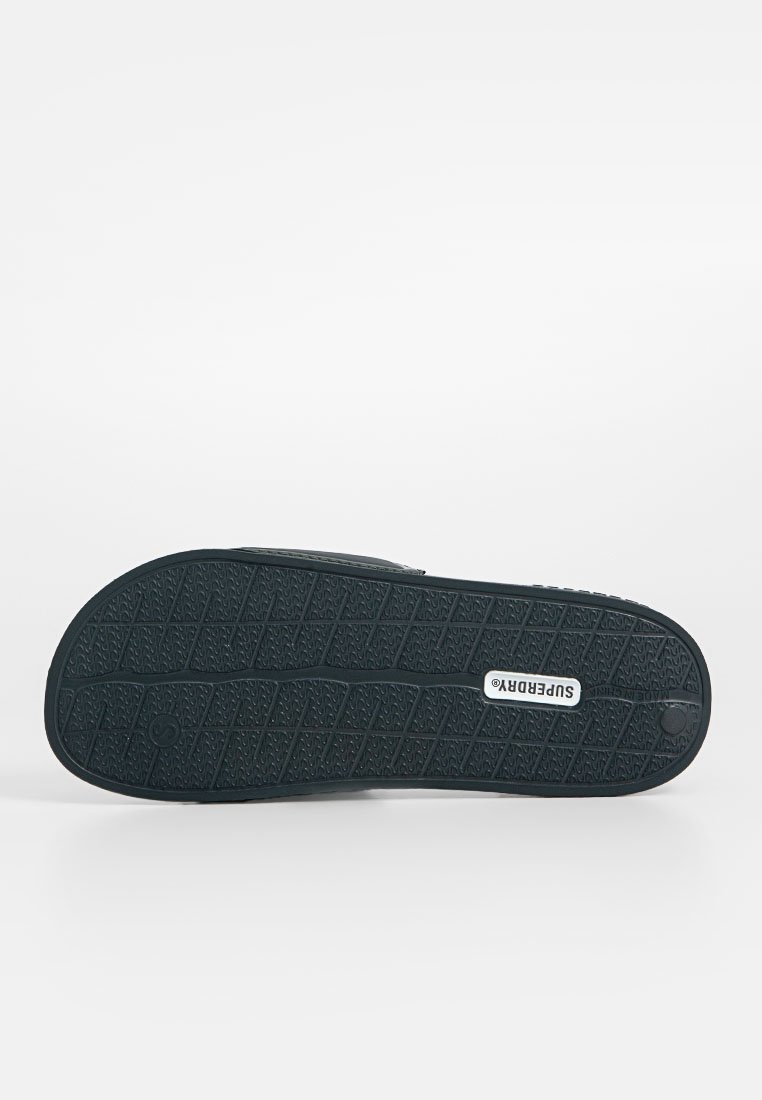 Semelle de chaussure en caoutchouc noir avec un motif texturé pour une meilleure adhérence et le logo "SUPERDRY" embossé. Design plat avec un bout arrondi.