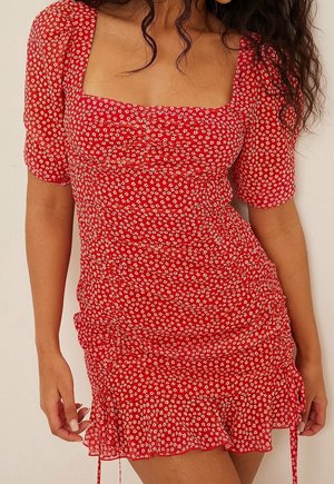 Mujer que lleva un vestido corto rojo con patrón floral blanco, mangas abullonadas, escote cuadrado y lados fruncidos con cordones.