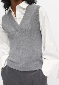 Gilet gris en maille avec un col en V, un côtes texturé et une coupe ajustée, superposé sur une chemise blanche à boutons avec de larges poignets.
