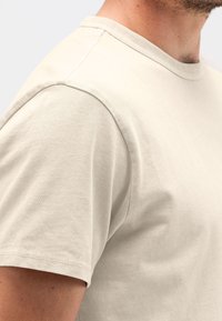Lichtbeige t-shirt met een ronde halslijn, korte mouwen en een zachte textuur. Zichtbare stiksels aan de mouwzoom en schoudernaad.