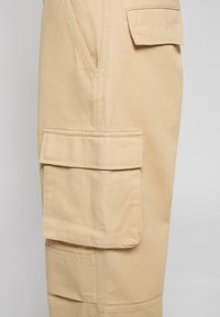 Pantalon cargo beige avec un tissu texturé, poches latérales et ourlet renforcé. Les détails de couture visibles améliorent la durabilité.