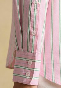 Gros plan sur une manche de chemise rose avec des rayures verticales vertes et bleues, présentant un poignet avec deux boutons blancs et des détails de boutonnières.