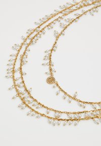 Gouden metalen ketting met meerdere strengen, versierd met kleine witte parels en decoratieve schakels, geaccentueerd met een bloemenhanger.