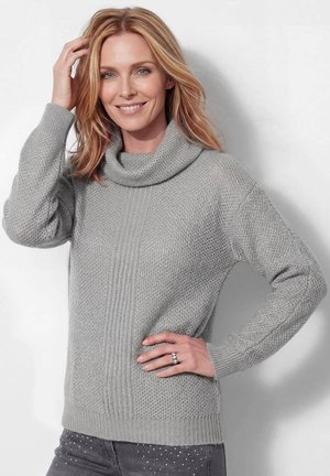 Vrouw met schouderlang blond haar lacht en draagt een lichtgrijze, gestructureerde gebreide trui en donkere broek met kleine witte stippen.