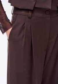 Brązowe spodnie na miarę z zakładkami, wykonane z gładkiego materiału. Posiadają boczną kieszeń oraz subtelny guzik przy talii.