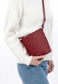 Bolso cruzado de cuero rojo con un acabado texturizado, forma cuadrada, correa ajustable y un discreto detalle de logo en la parte frontal.