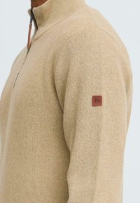 Beige gebreide trui met hoge kraag en ritssluiting. Heeft een gestructureerde stof en een bruin leren logo patch op de linkermouw.
