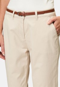 Pantaloni in cotone beige con una texture liscia, dotati di tasche laterali e una cintura in pelle marrone con una fibbia argentata per la regolazione.