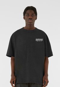 Sort oversized T-shirt lavet af bomuld, med et hvidt grafik på venstre forside af brystet. Korte ærmer og rund halsudskæring.