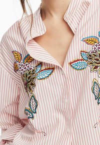 Gestreiftes Hemd in Pink und Weiß, mit farbenfroher Blumenstickerei in Blau, Grün und Lila, ausgestattet mit einem legeren Kragen und Knopfdetails.