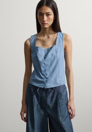 Femme aux longs cheveux foncés portant un haut sans manches bleu clair boutonné et un pantalon bleu foncé à cordon, sur un fond uni.