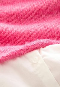 Maglione rosa lavorato a maglia con una texture morbida, sovrapposto a una camicia bianca con dettaglio dei bottoni.