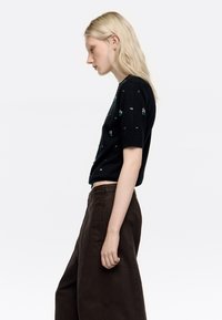 Suéter negro de manga corta con adornos verdes y plateados, combinado con pantalones marrones de corte amplio, con una textura suave y una alta cinturilla.