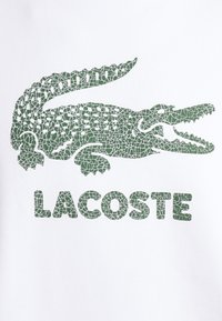 Camiseta blanca con un logotipo de cocodrilo verde texturizado y el texto "LACOSTE" debajo en el mismo color, transmitiendo un diseño informal.