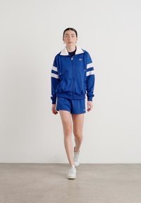 JAqueta atlética azul com riscas brancas e colarinho, combinada com calções correspondentes. Modelo usa ténis brancos. Tecido suave, design casual.