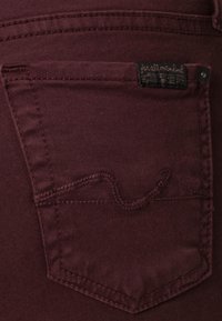 Los jeans de mezclilla burdeos presentan un bolsillo trasero con un patrón de olas bordado y una etiqueta con el texto "for all made kind".